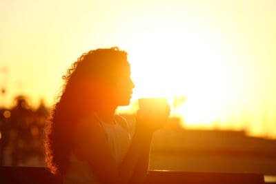 Side view of silhouette of a woman holding coffee mug sitting on a bench at sunset illustrating new possibilities open with soul retrieval.